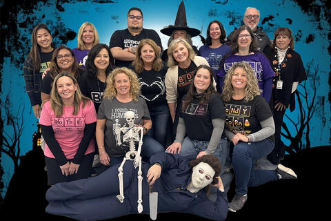 16 people dressed in periodic table shirts including person dressed as michael myers from the halloween movies with a skeleton sitting on top of them.  